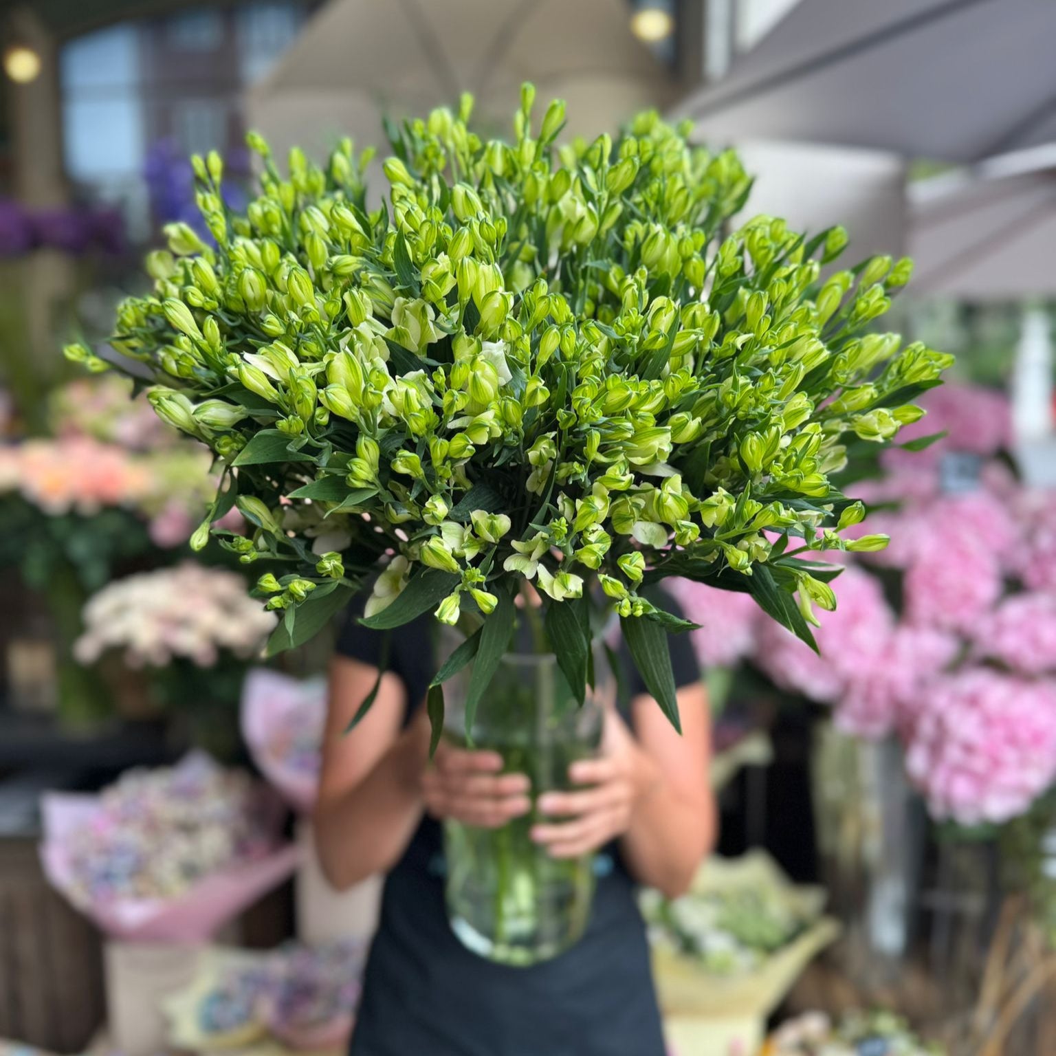 Person holding a bouquet of green flowers in a market setting with blurred flowers in the background