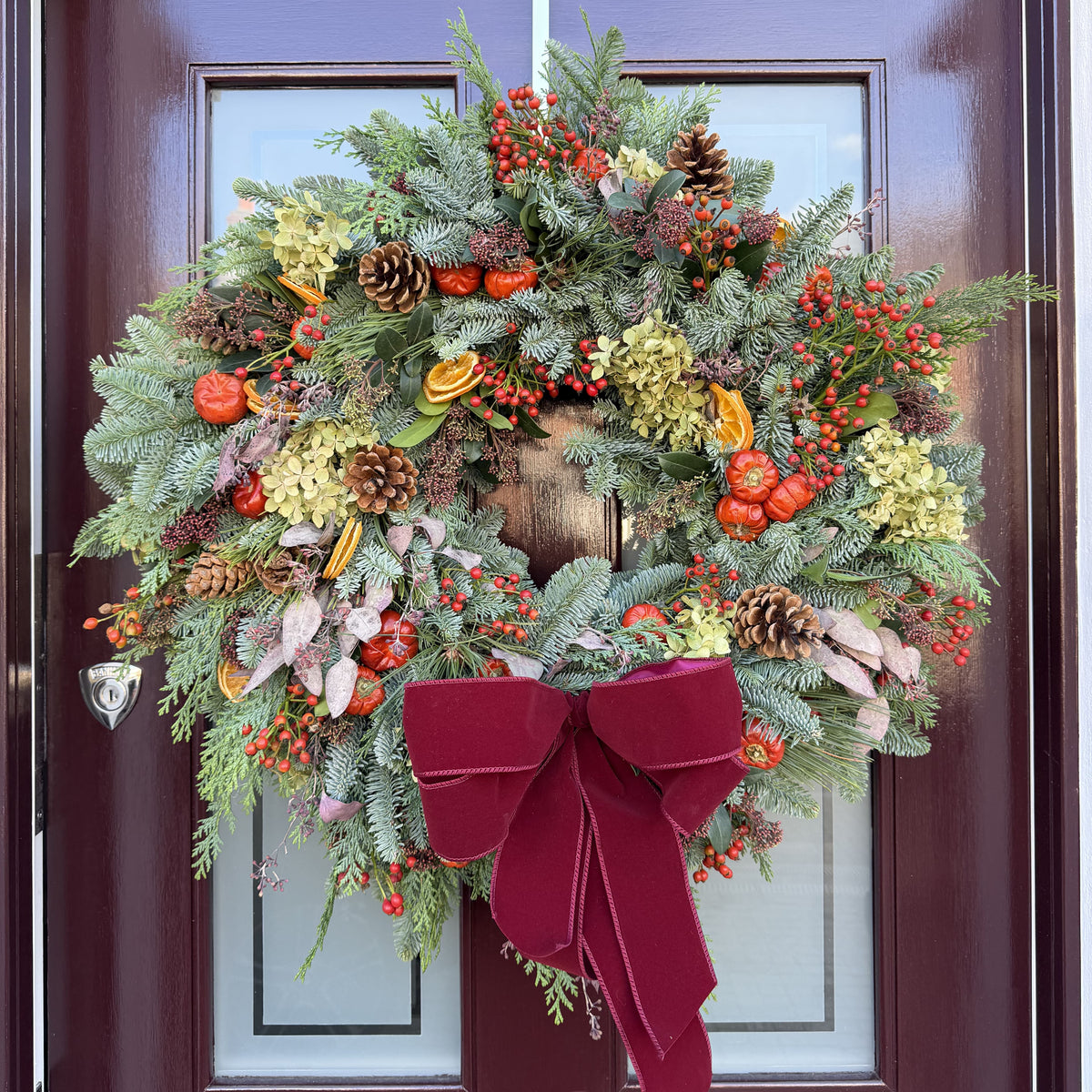 A festive Christmas wreath hanging on a dark reddish-brown front door. The wreath is made of green pine and fir branches with red berries, pinecones, small orange ornaments, dried flowers, and a large deep red velvet bow at the bottom.