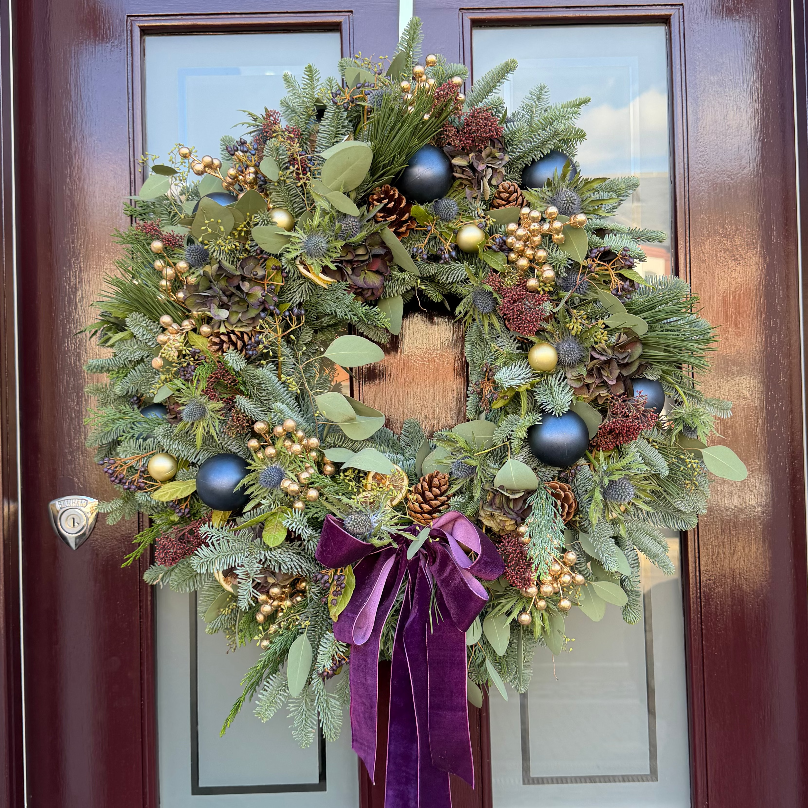 A festive Christmas wreath hanging on a dark reddish-brown front door. The wreath is decorated with green pine and fir branches, evergreen leaves, pinecones, small gold and red berries, blue ornaments, and a large purple velvet bow hanging below.